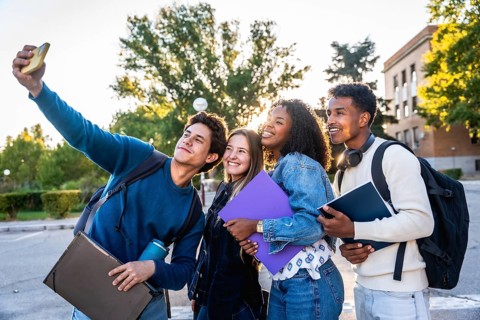 Diverse group of smiling university students recording a selfie outdoors on campus, holding books