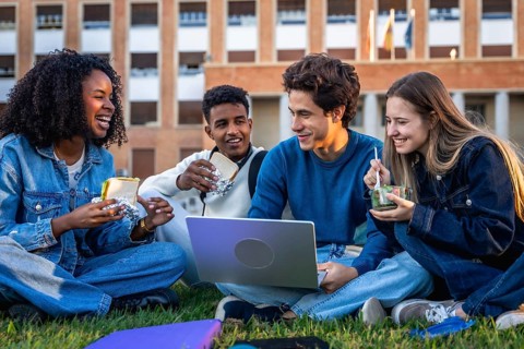 Group of happy young university students from different ethnicities eating, laughing, and using a