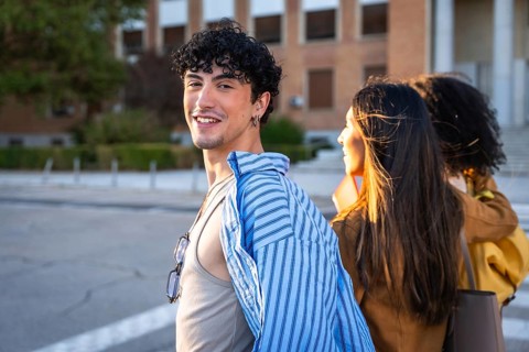 Young man with curly hair smiling at camera while walking with two female classmates on a sunlit