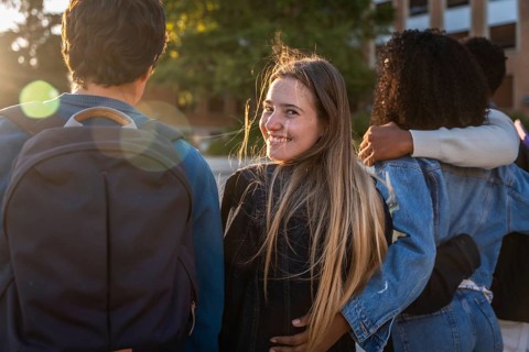 Group of young diverse university students walking arm in arm on campus, one woman turning to