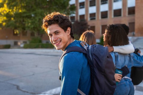 University student smiling at the camera with a backpack, walking with friends on campus, showing