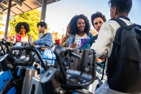 Young diverse students are talking and smiling at a public bicycle sharing station, using