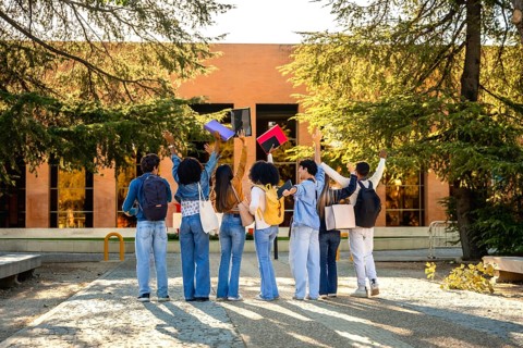 Diverse students seen from behind on a sunlit campus path throwing colorful notebooks into the air