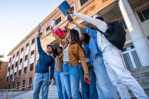 Group of happy young university students raising books and smartphones in the air, celebrating