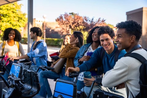 Group of smiling diverse university students standing at a bike sharing station on campus,