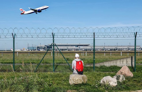 A man sits on a stone and watches the take-off of a British Airways plane through a barbed wire