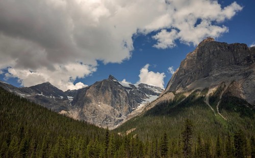 Rocky peaks of the Canadian Rocky Mountains under scattered clouds with a lush forest at the base