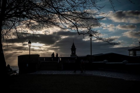 Figures silhouetted against sunset with dramatic clouds, tree, and lampposts in Helsingborg,