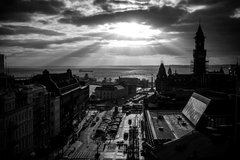 Black and white cityscape with dramatic clouds and sunlight over a harbor and buildings and looking