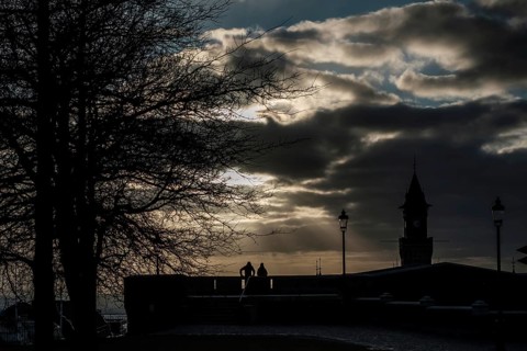 Silhouette scene with tree, lampposts, and tower against a dramatic cloudy sky at sunset in