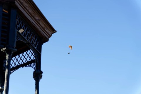 Paramotorist flying with a powered parachute against a bright clear blue sky, next to an old gazebo
