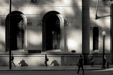 Black and white urban scene with people walking past a sunlit building casting strong shadows in