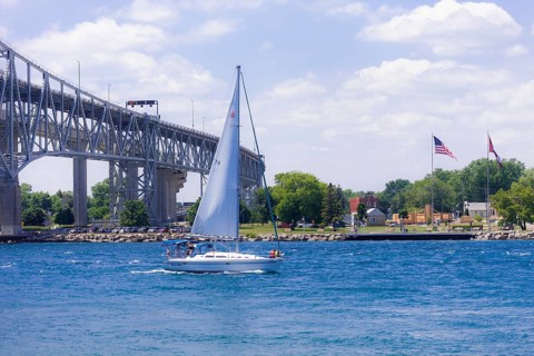 Sailboat gliding near the Blue Water Bridge, connecting Canada and the United States, with flags