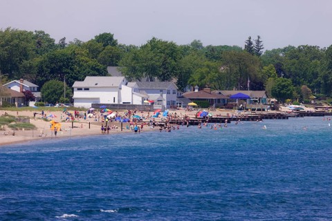 A busy beach scene with people enjoying the sun, umbrellas, and nearby houses along the St. Clair