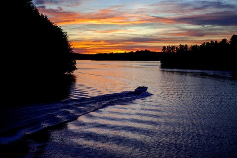 A boat moves across a lake during twilight, leaving ripples under a colorful sunset in Muskoka,