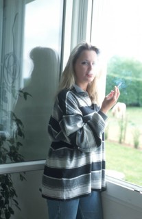 A woman with a striped shirt stands by a window, holding a cigarette, with natural light