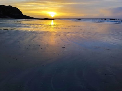 A serene sunset over a beach with calm ocean waters and rocks silhouetted against a golden sky