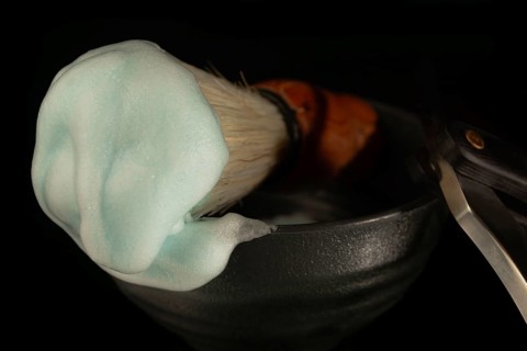 Shaving brush with teal lathered foam and straight razor in a dark bowl for men's grooming