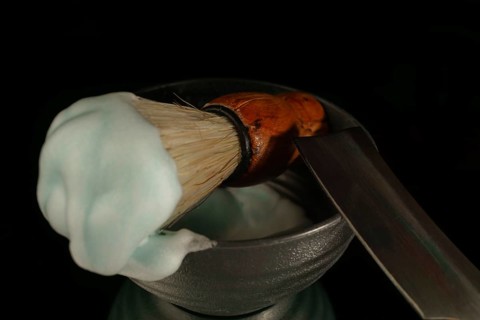 Shaving brush with cream and straight razor resting in a bowl on black background