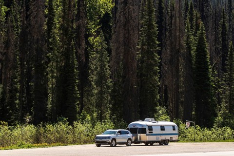 An Airstream travel trailer parked with a car by a dense forest, highlighting travel adventure in