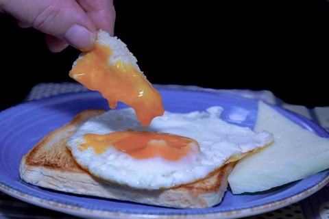 Close-up of a hand dipping bread into a runny egg yolk, served on toast with cheese, creating a