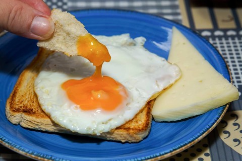 Close-up of dipping toast into a fried egg yolk, served with cheese on a blue plate, enjoying a