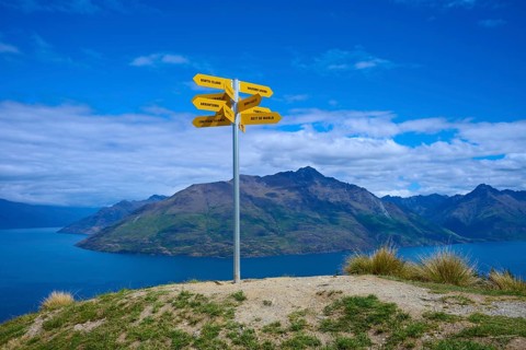 Yellow signposts on a hill in front of a vast mountain landscape under a blue sky, Queenstown,