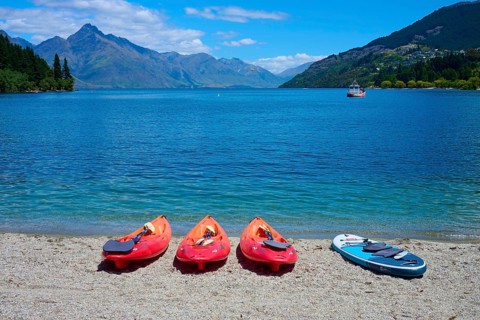 Red kayaks and paddleboard on the beach in front of a calm lake with mountain views, Queenstown,