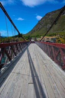 Wooden footbridge with red railing, shadows and mountain background, Kawarau Gorge Suspension
