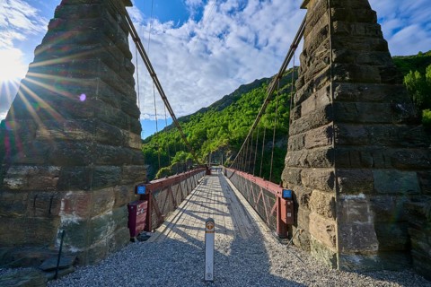 Old bridge with massive stone pillars surrounded by lush greenery under a bright blue sky, Kawarau