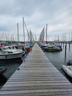 A long jetty runs through a harbour, surrounded by boats and masts on a calm, cloudy day, Laboe,
