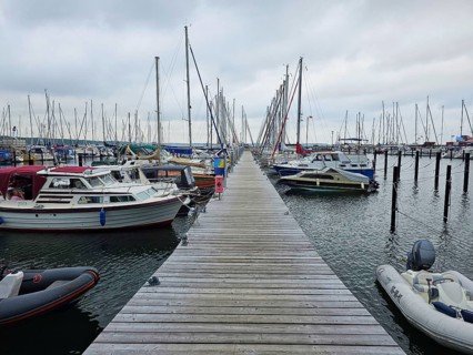 A wooden walkway leads through a harbour, lined with boats and masts under a cloudy sky, Laboe,