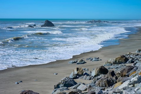 The waves gently lap against a wide sandy beach with scattered rocks, West Coast, South Island, New