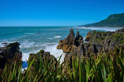 Barren cliffs and lush coastal vegetation meet the foaming sea under a clear sky, Pancake Rocks,