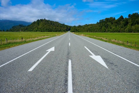 Long empty road with directional arrows through rural surroundings, West Coast, South Island, New