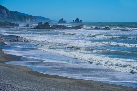 Dynamic waves roll onto a rocky beach under a bright blue sky, West Coast, South Island, New