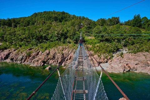 Suspension bridge over turquoise river, surrounded by dense forest under blue sky, Buller Gorge