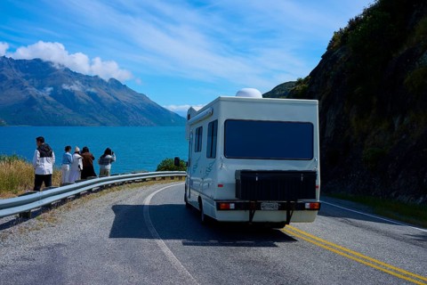 People enjoying travelling and a motorhome driving on a road along a lake surrounded by mountains,