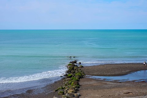 View of the calm sea with rocks leading into the water and a lone walker on the beach, Mana Bay,