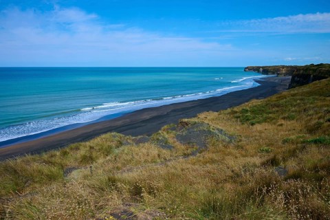 View of black sand beach with gentle waves, grassy coastline under clear sky, Mana Bay, Riverview