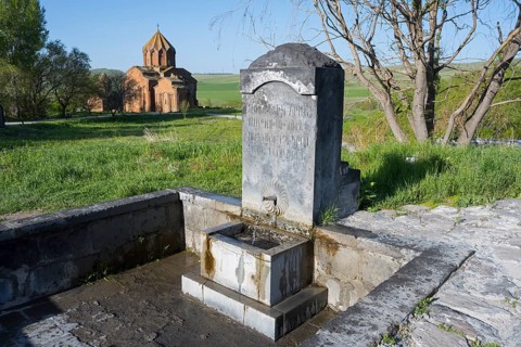 Stone fountain with engraving in front of a rural church in a green landscape, original spring,