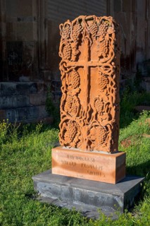 A detailed engraved red gravestone with a cross motif stands on a grassy area in the sunlight,
