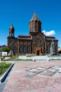 A red church with classical architecture and a stone monument under a clear sky, Church of the Holy