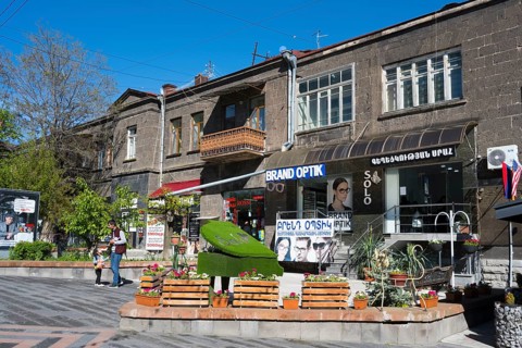 Pedestrian zone with optical shop and advertising signs, flanked by concrete buildings and trees,