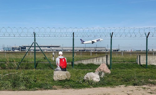 A man sits on a stone and watches an aircraft of the Polish airline LOT take off through a barbed