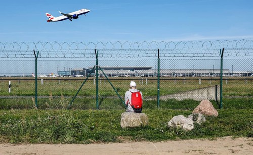 A man sits on a stone and watches the take-off of a British Airways plane through a barbed wire