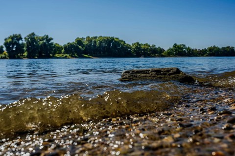 The sun shines on the calm waters of a river. Small waves gently lap against the shore, while the