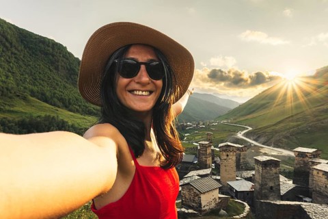 Smiling woman in hat and red dress taking selfie at sunset with medieval towers of Ushguli Georgia