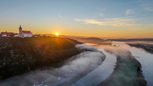 The sun rises over the Danube while mist drifts over the water. A tranquil landscape with trees and