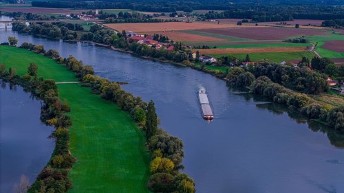 A cargo ship sails quietly on the Danube. The surroundings are green with fields and trees. The day
